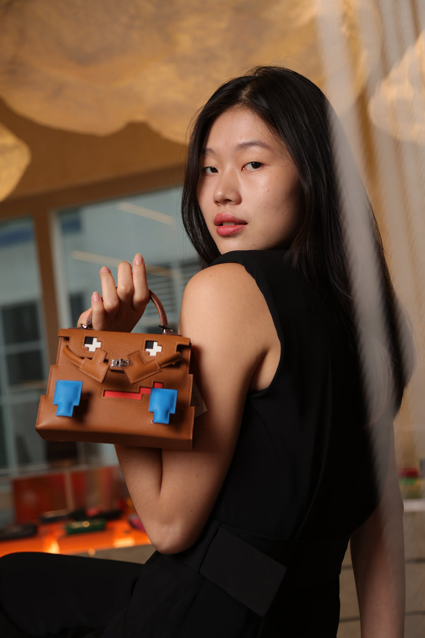 Model holding a brown Hermès Kelly Doll bag with playful blue and red details for The Orange Box, a luxury bag shop in BGC, Philippines.
