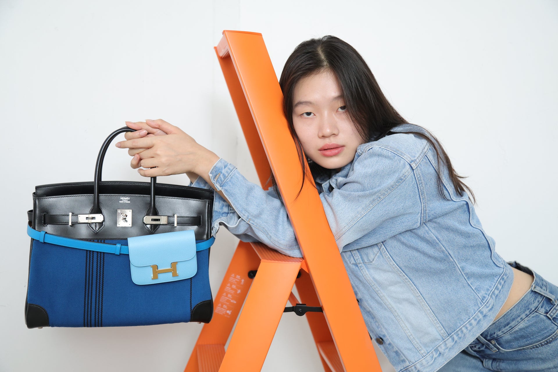 Model posing with a blue and black Hermès Birkin bag beside an orange ladder for The Orange Box, a luxury bag shop in BGC, Philippines.