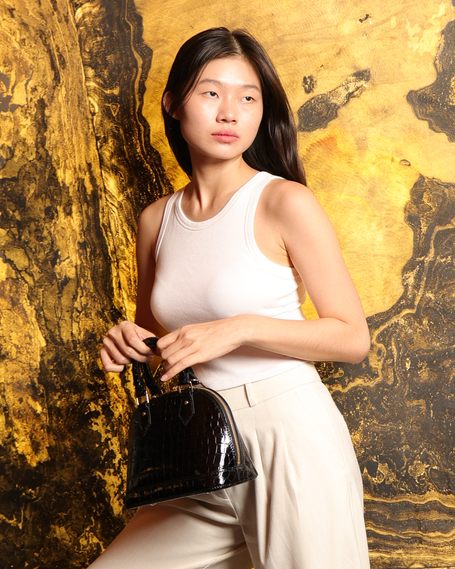 Model holding a LV black crocodile-embossed luxury handbag against a gold backdrop for The Orange Box, a luxury bag shop in BGC.